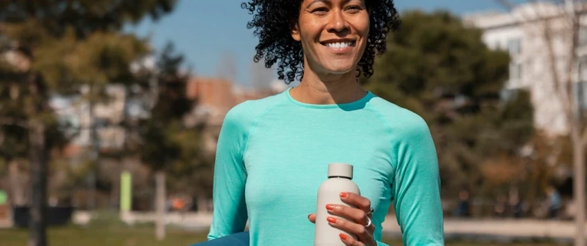 Woman holding a yoga mat and water bottle while building healthy habits outdoors