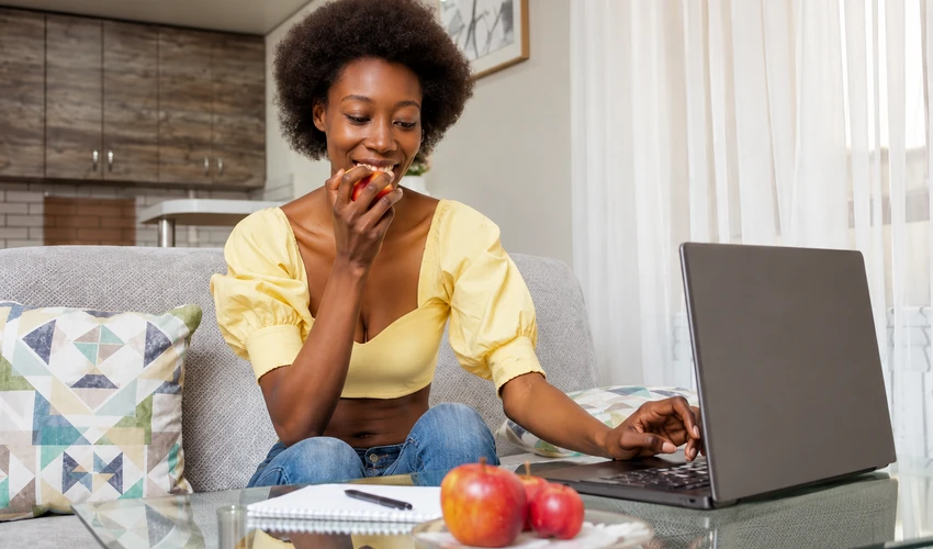 Woman eating an apple while working on a laptop at home